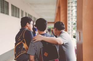 Two students embrace in a school hallway while a third stares out a window.