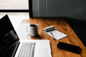 A laptop, coffee mug, cellphone, pen, and paper lying on top of a table