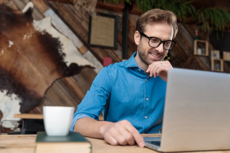 A smiling writer reviews copy on a laptop in a creative office setting with a book and a coffee mug nearby