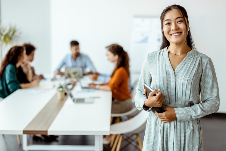 A smiling businessperson stands in front of colleagues meeting around a conference table.