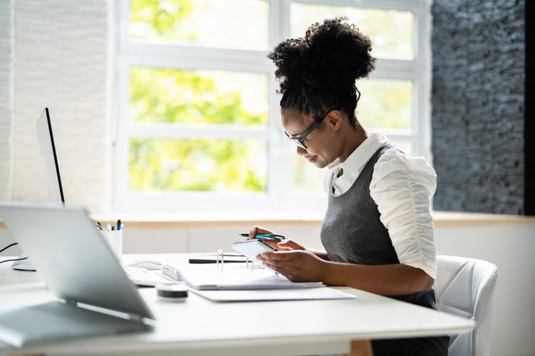 An accountant works at a desk with a laptop, using a calculator and writing on a notepad. 