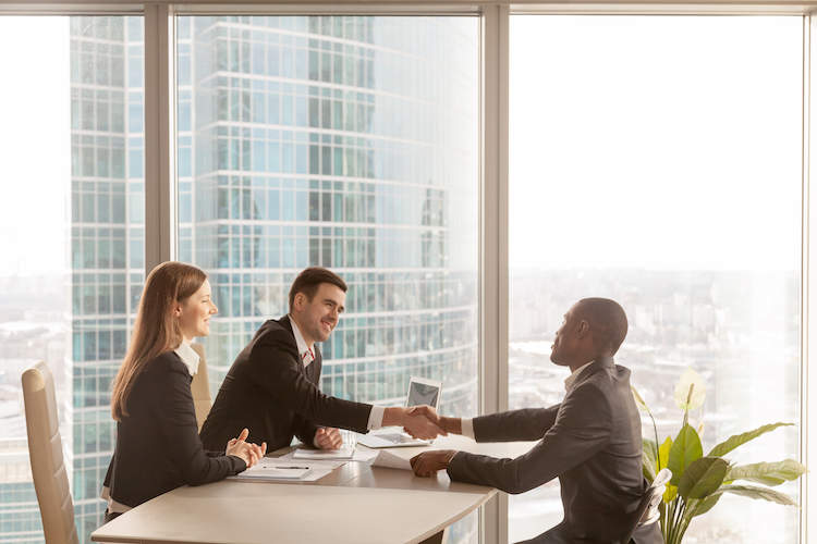 Friendly caucasian employers and confident african-american applicant handshaking during job interview sitting at office desk with big large window urban buildings cityscape at background, side view