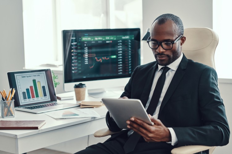 A data modeler consults a tablet while two monitors in the background display analytical graphs.