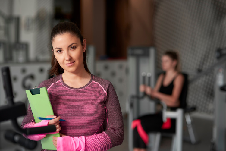 A fitness manager holding a clipboard stands in front of a client using a weight machine