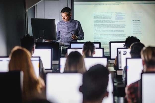 A professor shares their screen on a whiteboard while students work on computers.